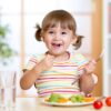toddler-aged girl with pigtails eating a plate of veggies with a huge smile on her face