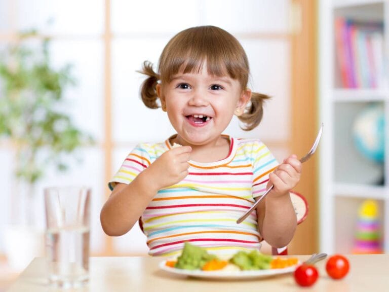 toddler-aged girl with pigtails eating a plate of veggies with a huge smile on her face
