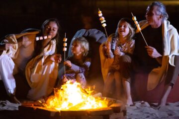 A family sitting around a campfire on the beach at night, roasting marshmallows on sticks and laughing together.