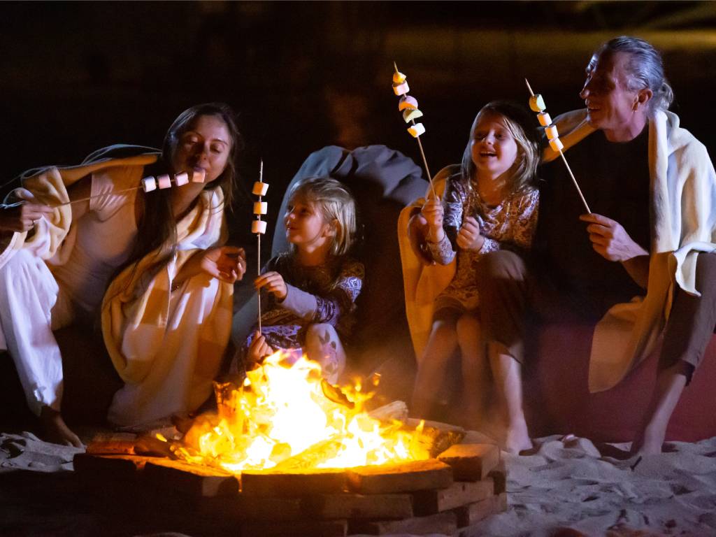 A family sitting around a campfire on the beach at night, roasting marshmallows on sticks and laughing together.