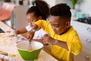 Two young children in a kitchen cooking. A little boy uses a whisk to beat eggs while a little girl forms dough out of flour.