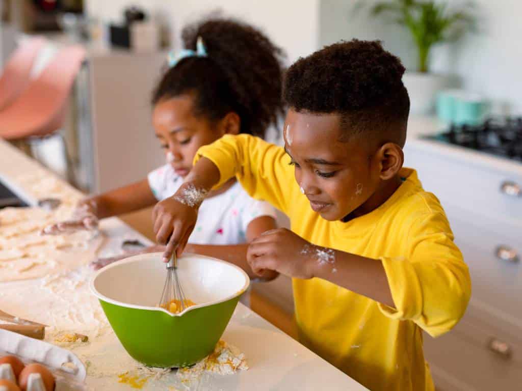 Two young children in a kitchen cooking. A little boy uses a whisk to beat eggs while a little girl forms dough out of flour.
