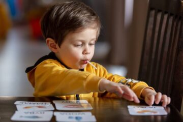 A young boy wearing a yellow hoodie, sitting at his desk reviewing his flash cards. He's enjoying a sucker and heavily focused.