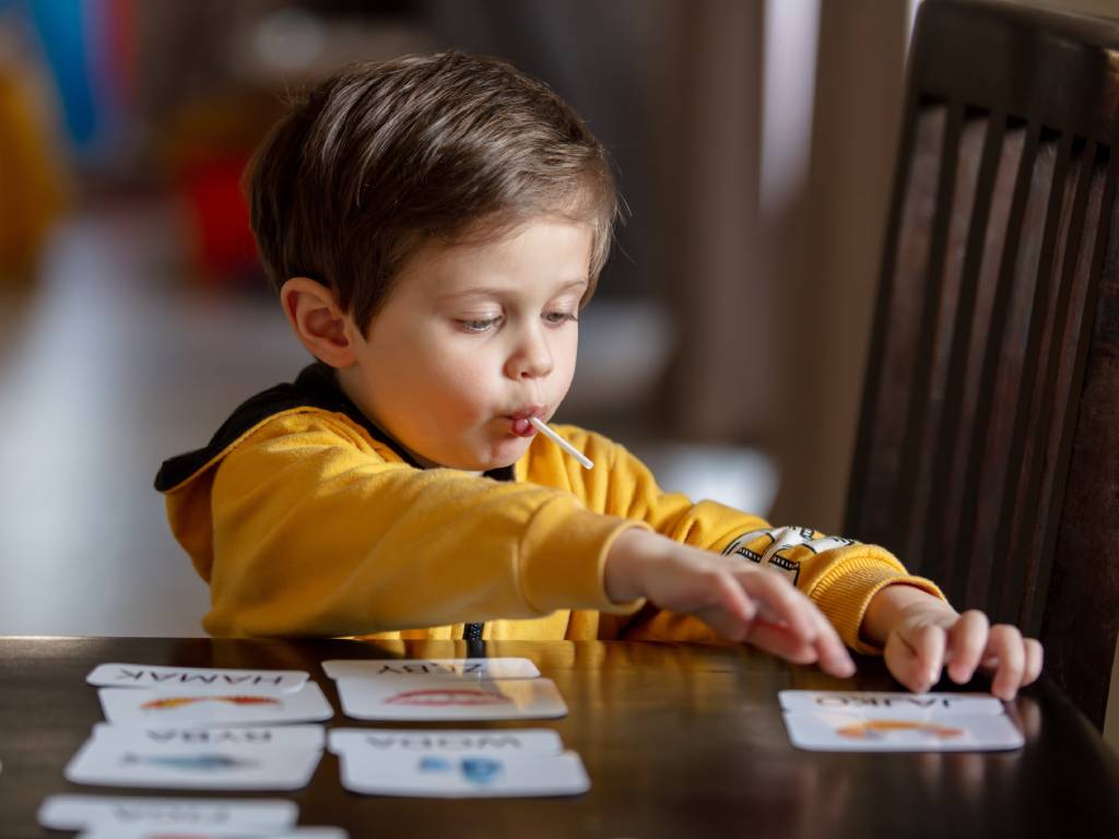 A young boy wearing a yellow hoodie, sitting at his desk reviewing his flash cards. He's enjoying a sucker and heavily focused.