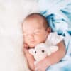 A sleeping newborn baby is wrapped in a soft blue security blanket and hugs a cute stuffed lamb lovey.