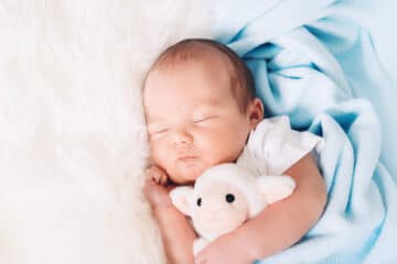 A sleeping newborn baby is wrapped in a soft blue security blanket and hugs a cute stuffed lamb lovey.