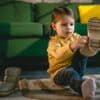 A young toddler wearing a yellow sweater and blue jeans is putting on her boots at home. She's in front of a green sofa.