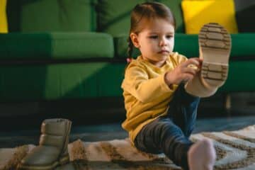 A young toddler wearing a yellow sweater and blue jeans is putting on her boots at home. She's in front of a green sofa.