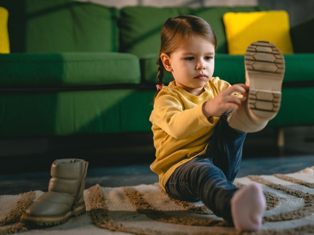 A young toddler wearing a yellow sweater and blue jeans is putting on her boots at home. She's in front of a green sofa.