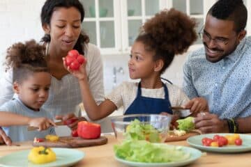 A happy young family with two little kids prepare a healthy salad by chopping up lettuce, tomatoes, and peppers.