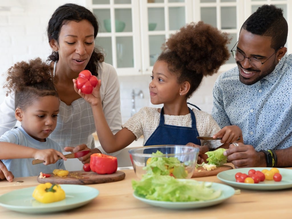 A happy young family with two little kids prepare a healthy salad by chopping up lettuce, tomatoes, and peppers.