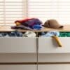 An annoying pile of clutter on the dresser with the drawers open and items falling out. Clothing and a hat are piling up.