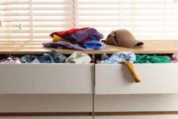 An annoying pile of clutter on the dresser with the drawers open and items falling out. Clothing and a hat are piling up.