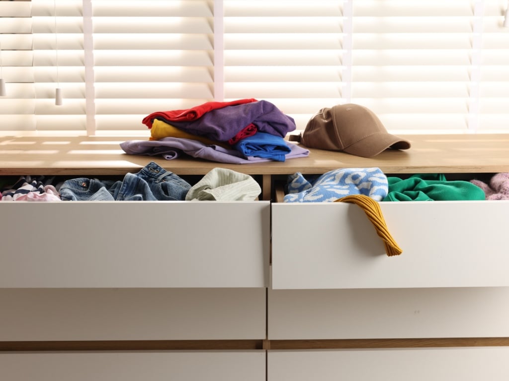 An annoying pile of clutter on the dresser with the drawers open and items falling out. Clothing and a hat are piling up.