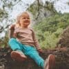 A young child sitting on a log in nature and smiling goofily at the camera as they enjoy the snack in their hand.