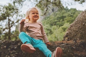 A young child sitting on a log in nature and smiling goofily at the camera as they enjoy the snack in their hand.