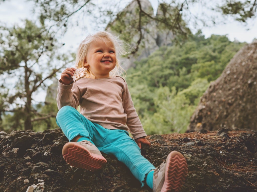 A young child sitting on a log in nature and smiling goofily at the camera as they enjoy the snack in their hand.