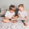 A young girl and a young boy sit on a bed laughing while looking at a book. Two pillows sit behind the kids.