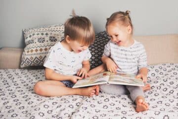 A young girl and a young boy sit on a bed laughing while looking at a book. Two pillows sit behind the kids.