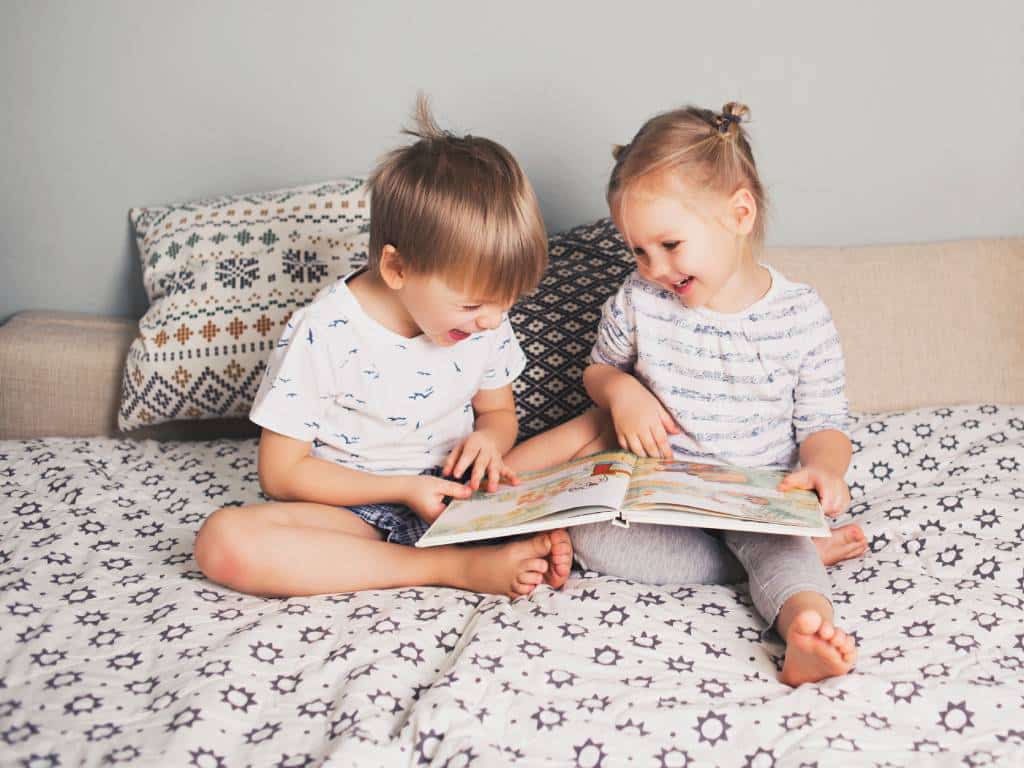 A young girl and a young boy sit on a bed laughing while looking at a book. Two pillows sit behind the kids.