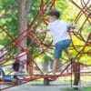 A little boy in a white shirt, blue shorts, and blue sandals climbs on red climbing ropes at the playground.