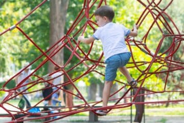 A little boy in a white shirt, blue shorts, and blue sandals climbs on red climbing ropes at the playground.