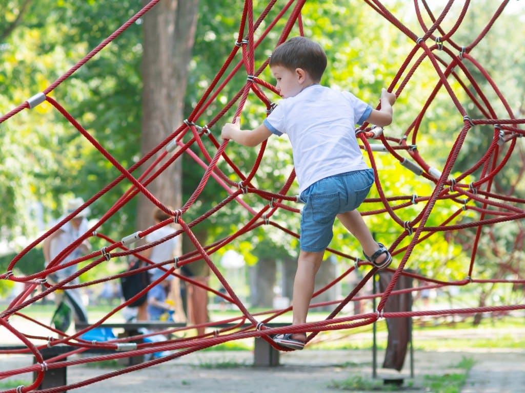 A little boy in a white shirt, blue shorts, and blue sandals climbs on red climbing ropes at the playground.