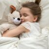 A little girl sleeps happily as she cuddles with her stuffed dog and lays on a cozy bed with cream-colored sheets.