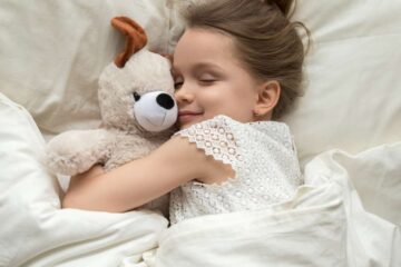 A little girl sleeps happily as she cuddles with her stuffed dog and lays on a cozy bed with cream-colored sheets.