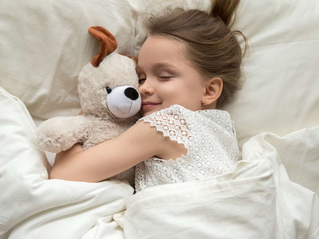 A little girl sleeps happily as she cuddles with her stuffed dog and lays on a cozy bed with cream-colored sheets.