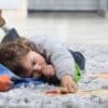 A toddler laying on the floor playing with a few toys and sucking his thumb. He's using his other hand to work on a puzzle.