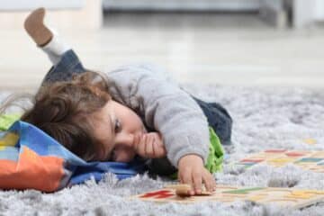 A toddler laying on the floor playing with a few toys and sucking his thumb. He's using his other hand to work on a puzzle.