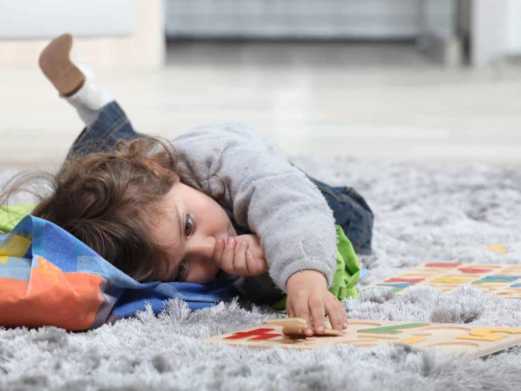 A toddler laying on the floor playing with a few toys and sucking his thumb. He's using his other hand to work on a puzzle.