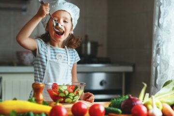 A happy child girl prepares a vegetable salad in the kitchen. She's eating a tomato and cucumber salad with a serving spoon.