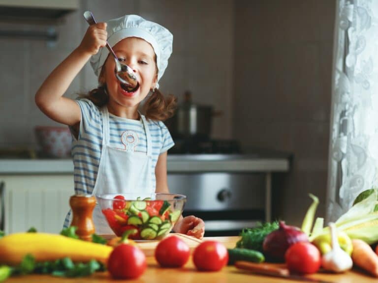 A happy child girl prepares a vegetable salad in the kitchen. She's eating a tomato and cucumber salad with a serving spoon.