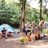 A large family with all ages gathered around a food platter situated on a log in a forested campground.