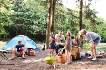 A large family with all ages gathered around a food platter situated on a log in a forested campground.