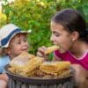 Two young girls are sitting on the grass outside in front of a large plate of honeycomb as one child eats a big piece.