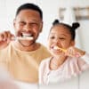 A Black father and his daughter standing in the bathroom vanity mirror brushing their teeth together.