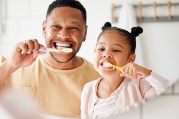 A Black father and his daughter standing in the bathroom vanity mirror brushing their teeth together.