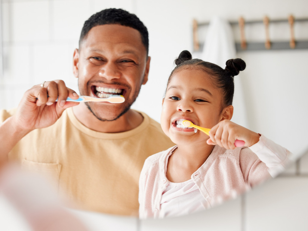 A Black father and his daughter standing in the bathroom vanity mirror brushing their teeth together.