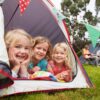 Three little girls lying at the door of a camping tent and smiling. Their parents sit in camping chairs in the background.