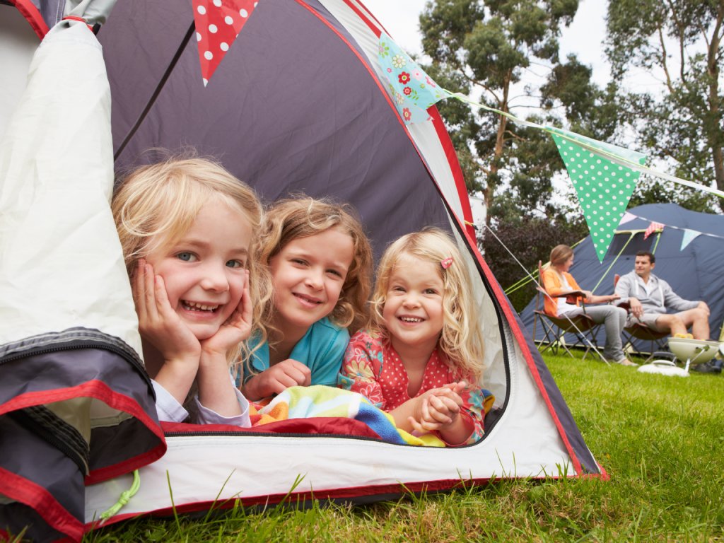 Three little girls lying at the door of a camping tent and smiling. Their parents sit in camping chairs in the background.