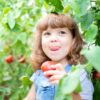 A young girl standing by a row of tomato bushes, holding a tomato in one hand while licking her upper lip.