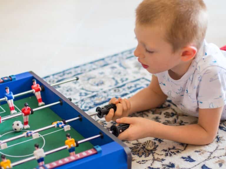A young boy laying on the floor with a rug beneath him. He holds the rods of a blue mini foosball table.
