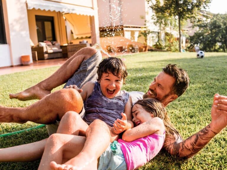 A man and two kids are laughing and playing in a backyard with a nearby hose sprinkling water on them.