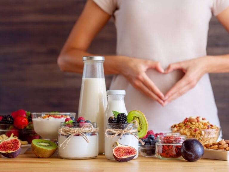 A spread of gut-friendly foods, including fruits, yogurt, and nuts, in front of a woman making a heart over her stomach.
