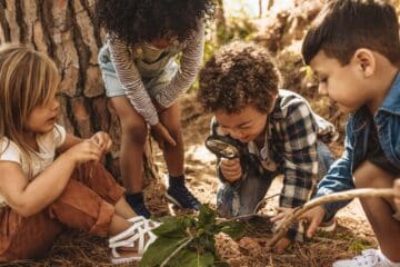 Four kids gathered around a cluster of green leaves in a forest. One holds a stick and another holds a magnifying glass.