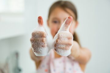 A young girl in a pink shirt hold out two thumbs up. Her hands are covered in suds.