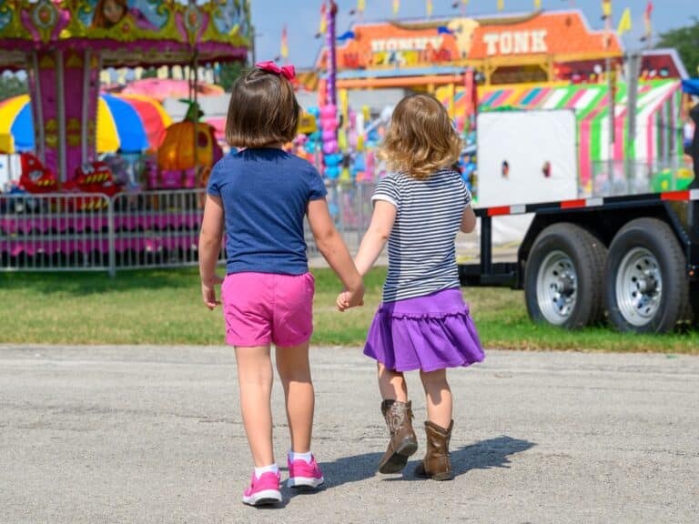 Two young girls hold hands and walk toward a county fair. Carnival rides are visible in the background.